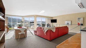 A modern living room with a red sofa, a dining area with a white table, and large windows offering a view of the outdoors.