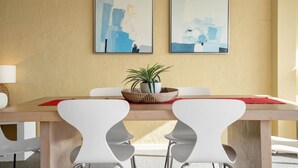 A dining area with a wooden table, white chairs, two abstract paintings, a potted plant, and a red placemat.