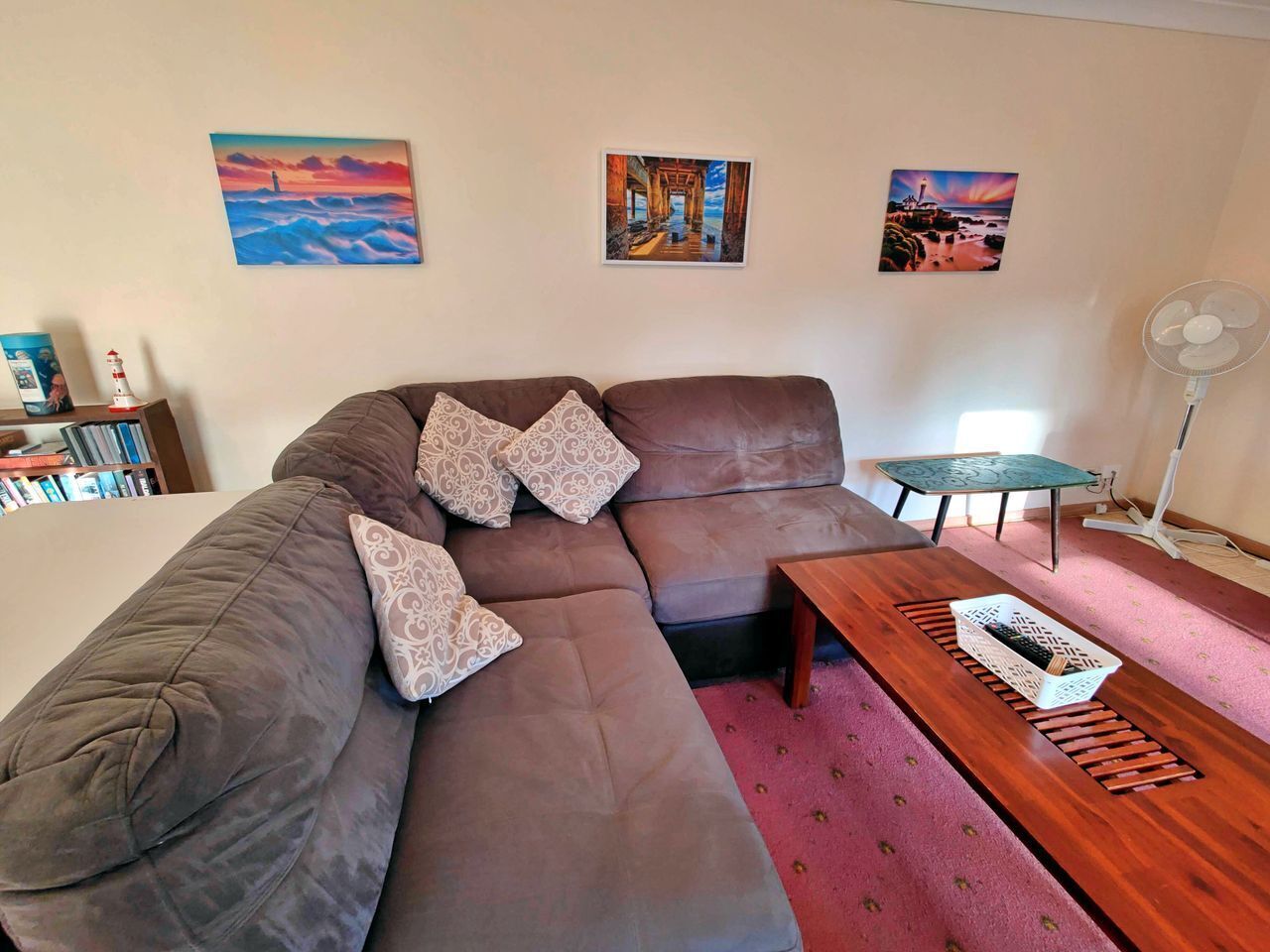 Ein Wohnzimmer mit einem Ecksofa, einem Couchtisch und einem kleinen Tisch mit Ventilator. Drei gerahmte Bilder hängen an der Wand.