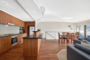 A modern kitchen with wooden cabinets, a central island, and a dining area with a view. - Butter Factory 9 (Apollo Bay)