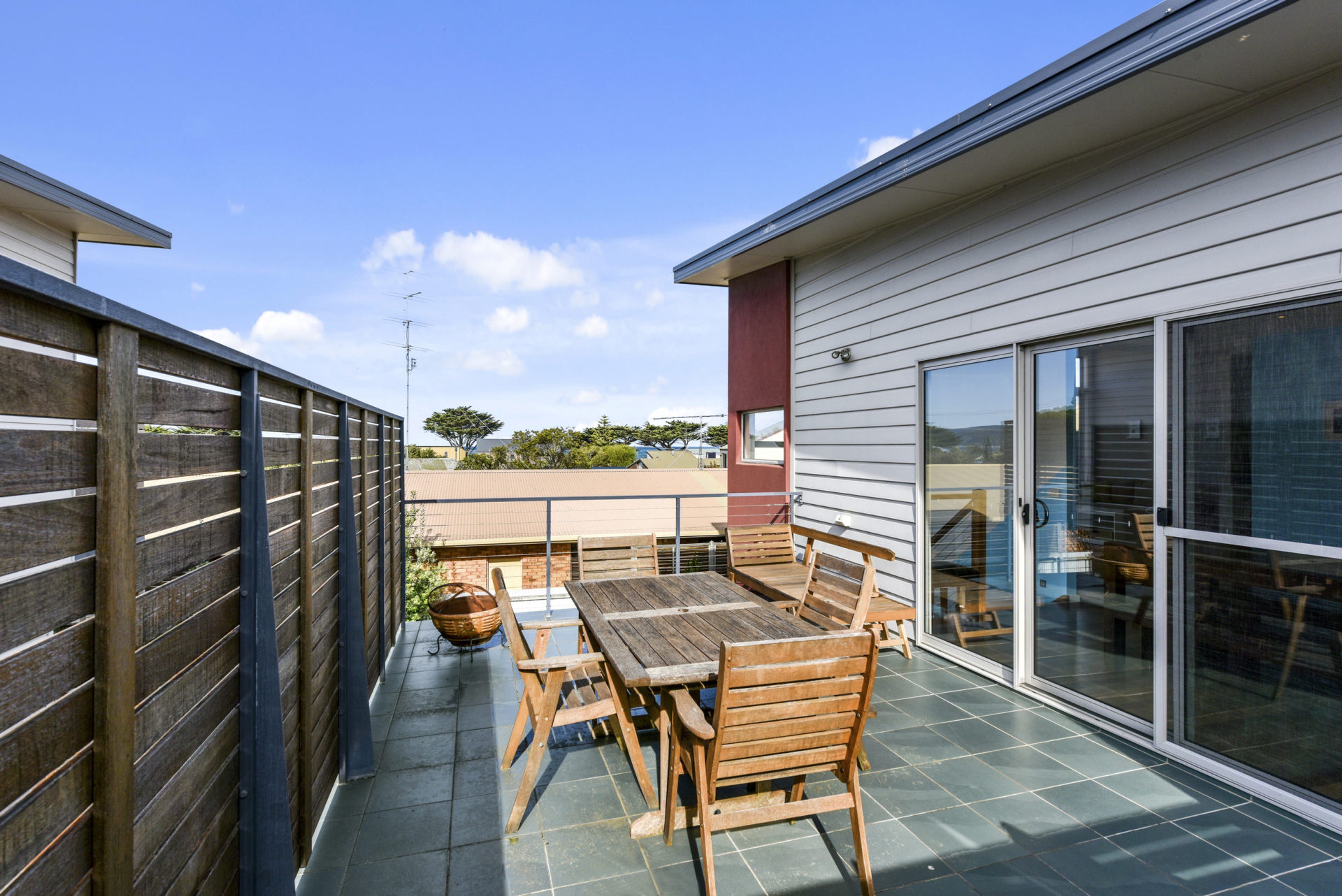 Une terrasse avec une table et des chaises en bois, une grande fenêtre et une vue sur un bâtiment en arrière-plan.