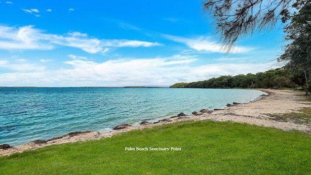 Une plage avec une eau d’un bleu limpide, un espace herbeux au premier plan et une zone boisée en arrière-plan.