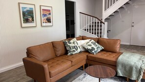 A brown leather sectional sofa with floral cushions, a round wooden coffee table, and a patterned throw blanket.