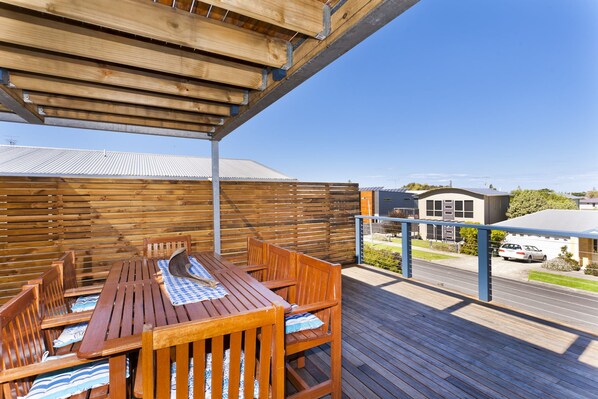 An outdoor wooden deck with a table and chairs, a covered area, and a view of a residential building in the background.