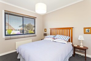 A bedroom with a bed, a bedside table, a lamp, a window with blinds, and a picture frame on the wall.