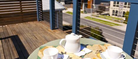 Uma mesa com bule, xícaras, pires e cupcakes em um terraço com vista para uma rua e edifícios.