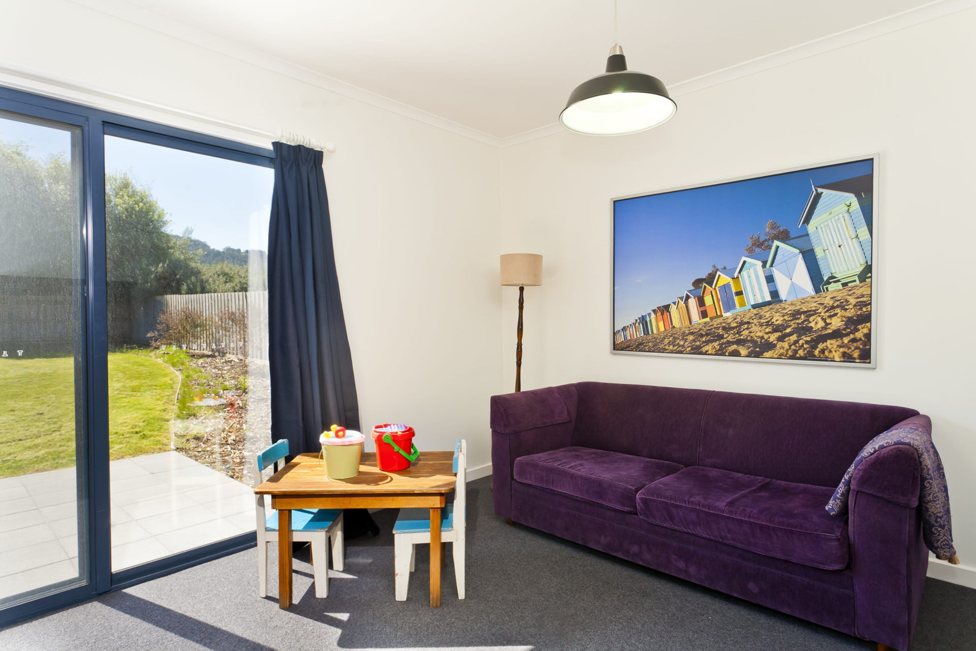 A room with a purple sofa, a dining table with chairs, a lamp, and a large window with a view of beach huts.