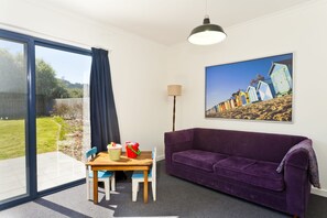 A room with a purple sofa, a dining table with chairs, a lamp, and a large window with a view of beach huts.
