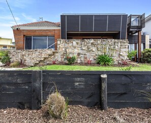 A modern house with a stone wall, a wooden deck, and a garden with various plants. - Absolute Beachfront Views Apollo Bay (Apollo Bay)