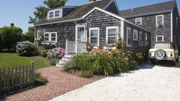 A two-story house with a front porch, a vehicle parked in the driveway, and a variety of flowers in the garden. - Renovated Cottage off Main Street- Great Location (Nantucket)