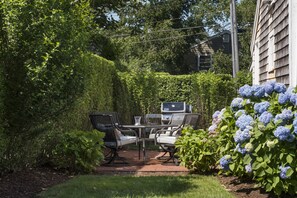 An outdoor patio with a table and chairs, surrounded by lush greenery and hydrangea flowers. - Renovated Cottage off Main Street- Great Location (Nantucket)
