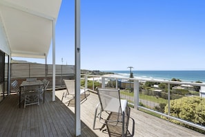 A wooden deck with a table and chairs, overlooking a coastal view with buildings and a clear sky.