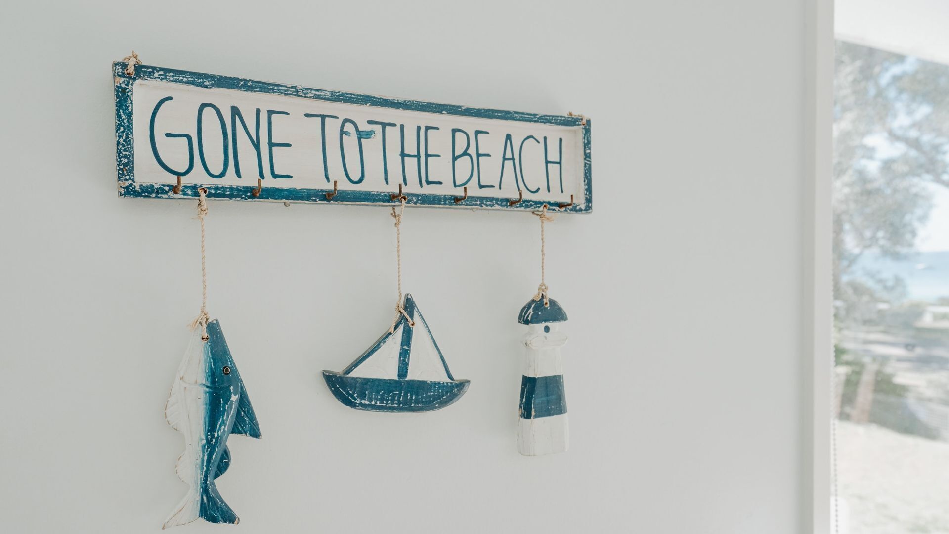 A wall with a sign that reads "GONE TO THE BEACH" and three hanging items: a sailboat, a piece of clothing, and a lighthouse.