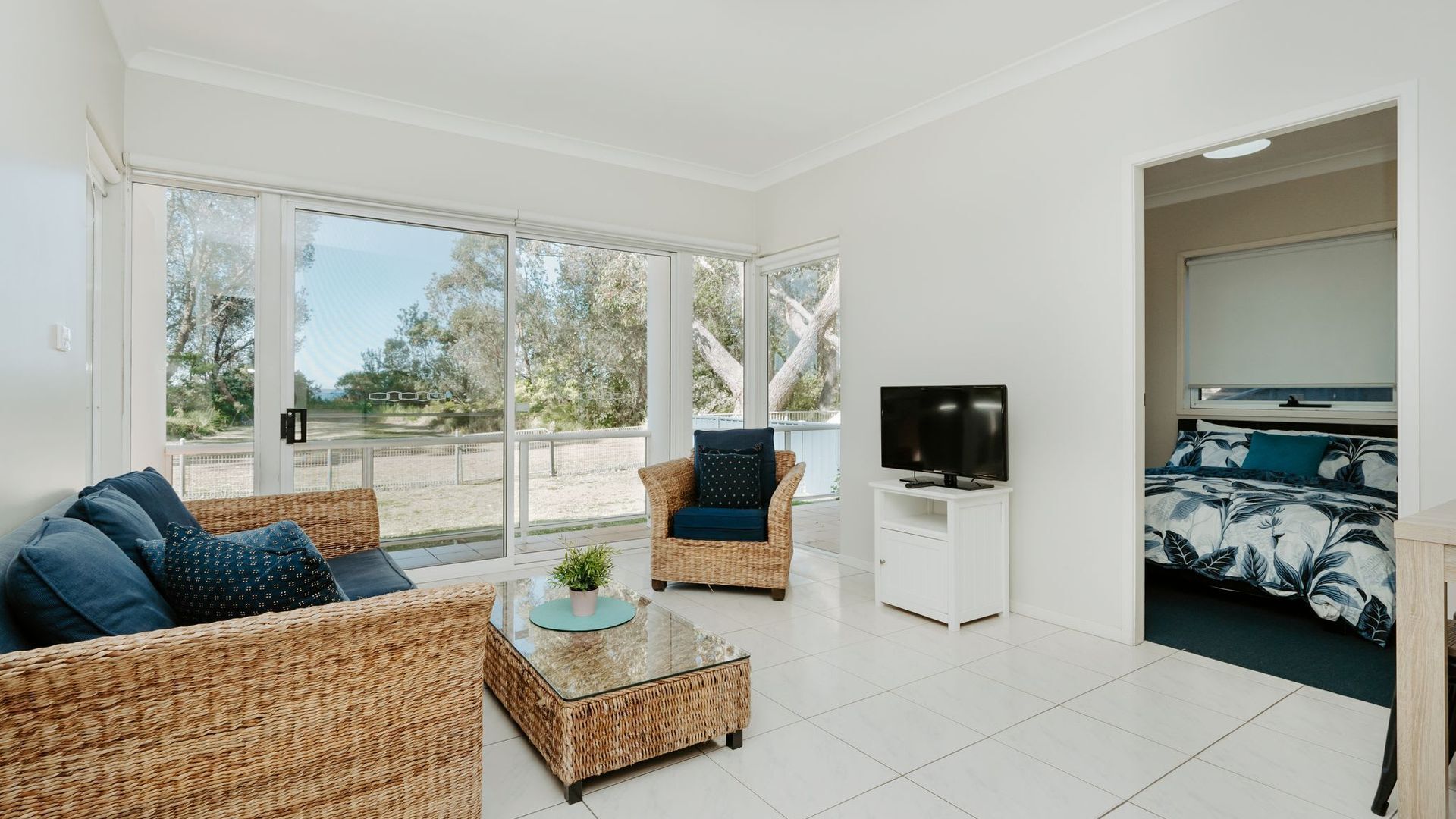 A spacious living area with wicker furniture, a glass coffee table, a flat-screen TV, and a bed with a floral bedspread.
