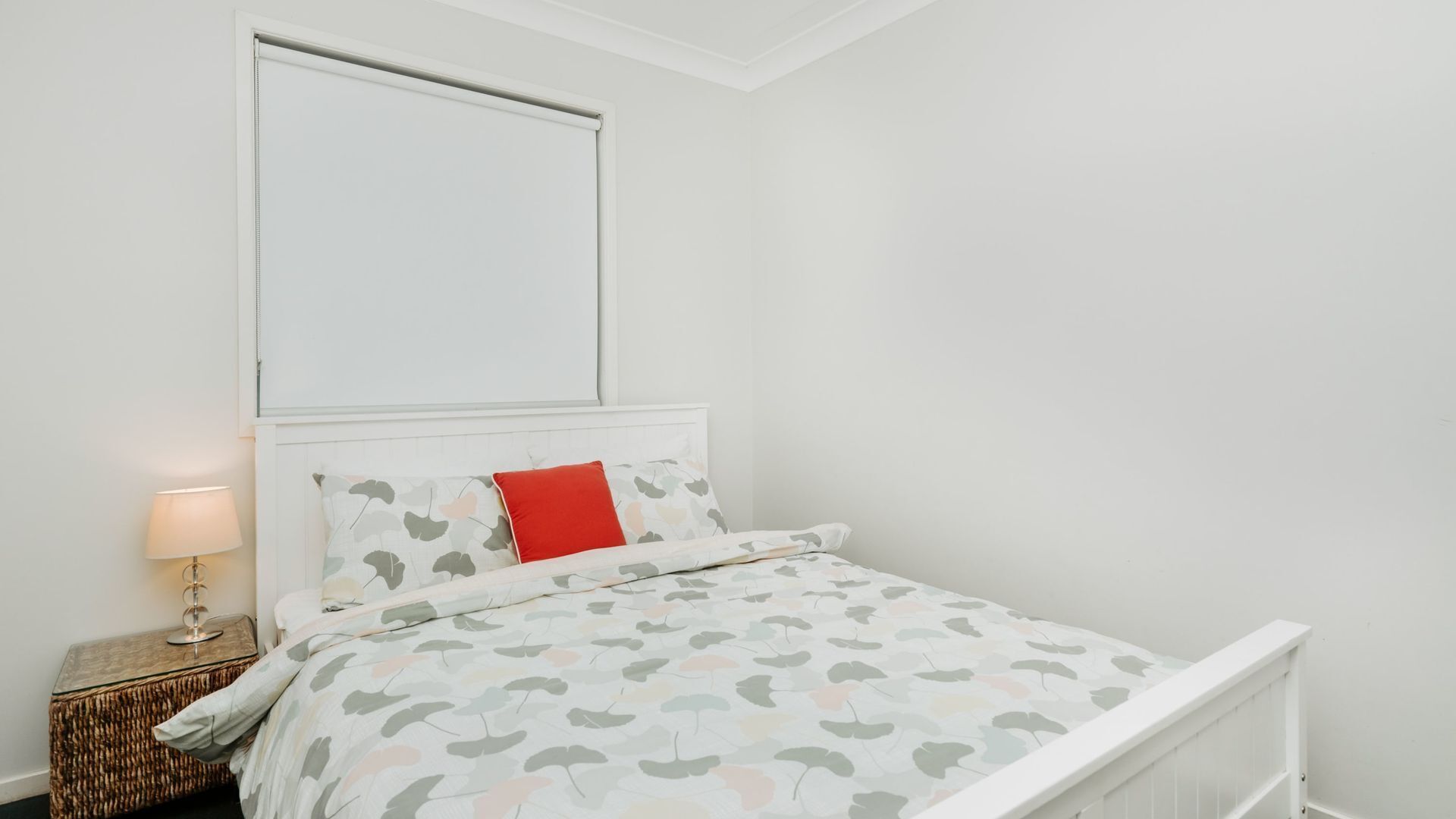 Uma cama bem arrumada com colcha estampada, um travesseiro vermelho e uma luminária de cabeceira em uma mesa de madeira.