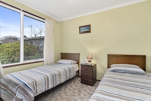 A bedroom with two single beds, a nightstand, a lamp, and a window with a view of trees and a building. - Low (Apollo Bay)