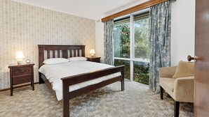 A bedroom with a large bed, a nightstand, a lamp, a window with curtains, and a view of greenery outside.