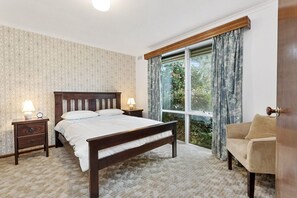 A bedroom with a large bed, a nightstand, a lamp, a window with curtains, and a view of greenery outside.