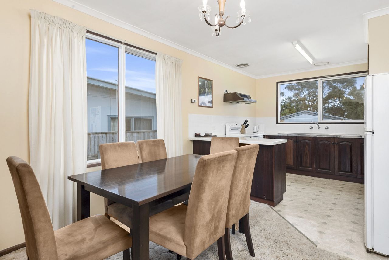 A dining area with a dark wooden table and beige upholstered chairs, a kitchen with dark wooden cabinets, and a large window with a view of a house.