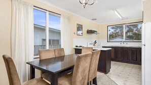 A dining area with a dark wooden table and beige upholstered chairs, a kitchen with dark wooden cabinets, and a large window with a view of a house.
