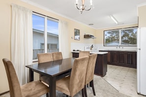 A dining area with a dark wooden table and beige upholstered chairs, a kitchen with dark wooden cabinets, and a large window with a view of a house.