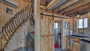 Un intérieur rustique avec un escalier en bois, un coin cuisine doté d’un évier et de placards, et un espace repas avec des tabourets.