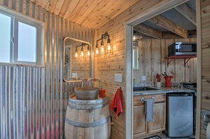 A rustic kitchen with wooden walls, a wooden barrel sink, a stainless steel refrigerator, and a microwave on a shelf.