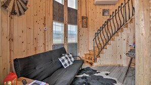 Un intérieur de chalet chaleureux, avec un revêtement mural en bois, un canapé noir agrémenté de coussins à carreaux, un tapis en peau de vache, une décoration représentant un moulin à vent et un escalier en colimaçon.