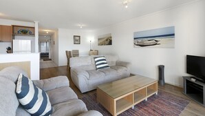 Ein modernes Wohnzimmer mit einer Couch, einem Couchtisch und einem Flachbildfernseher. An der Wand hängt ein Gemälde mit einer Strandansicht.