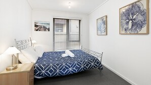 Ein Schlafzimmer mit einem Bett, einem Nachttisch, einer Lampe, einem Fenster mit Jalousien und Wandbildern.