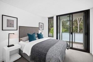A bedroom with a bed, a nightstand, a lamp, and a view through a sliding glass door to an outdoor balcony.