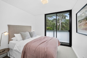 A bedroom with a large bed, a bedside table, a floor lamp, and a view of the outdoors through a sliding door.