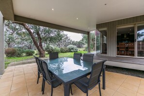 A covered patio with a glass-top table and chairs, surrounded by a garden with trees and shrubs. - Haven On Harvey (Anglesea)