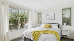 A bedroom with a large window, a bed with white bedding and a yellow throw, a bedside table with a lamp, and a framed picture on the wall.