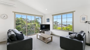 A modern living room with two sofas, a coffee table, a TV, and large windows with a view of greenery and the sea.