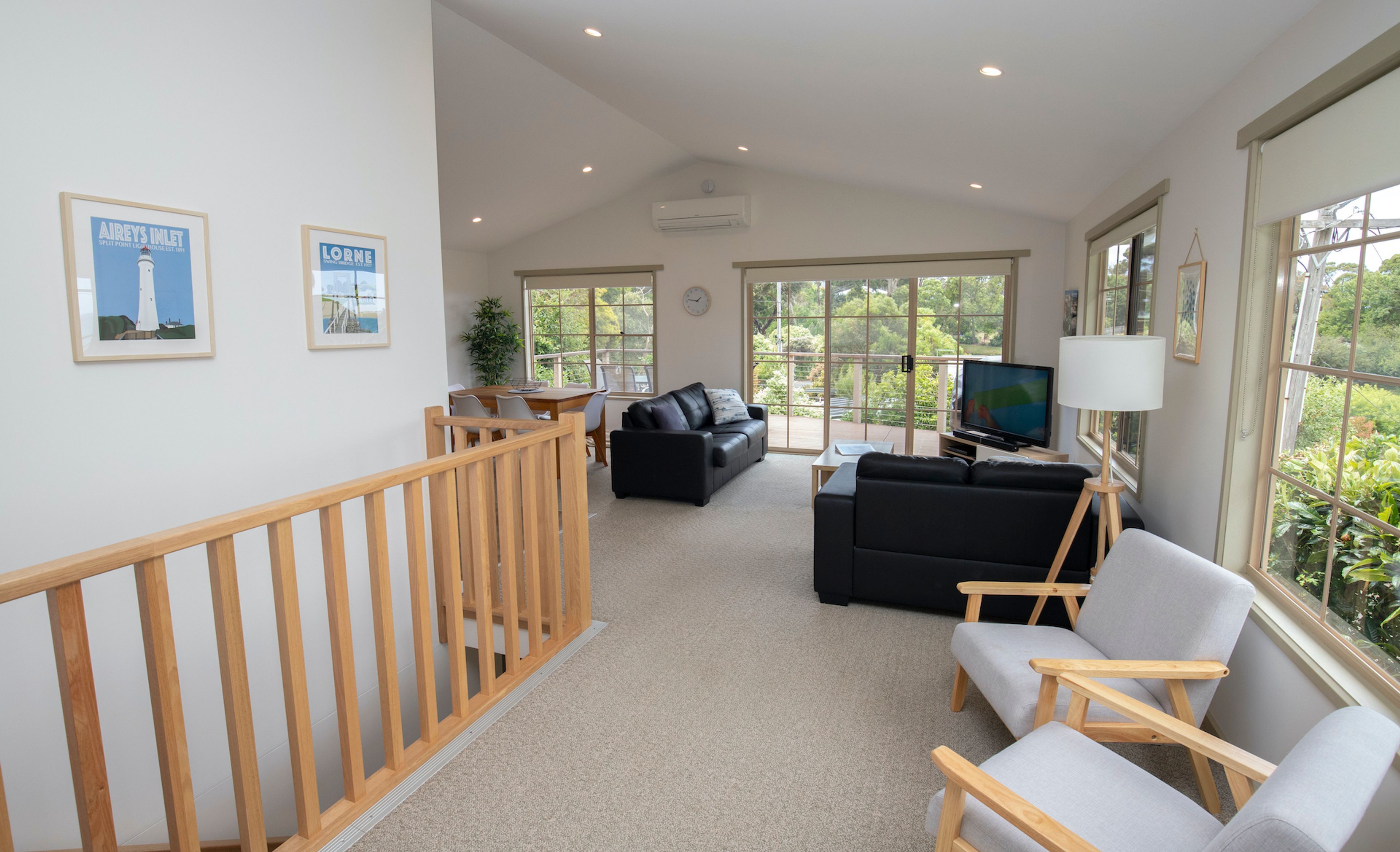 A spacious living area with a wooden staircase, a black leather sofa, a grey armchair, a television, and a large window with a view of greenery.