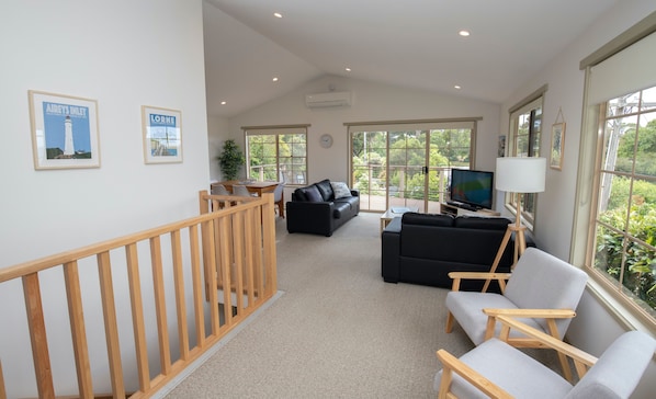 A spacious living area with a wooden staircase, a black leather sofa, a grey armchair, a television, and a large window with a view of greenery.