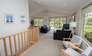 A spacious living area with a wooden staircase, a black leather sofa, a grey armchair, a television, and a large window with a view of greenery. - Vardon (Lorne)