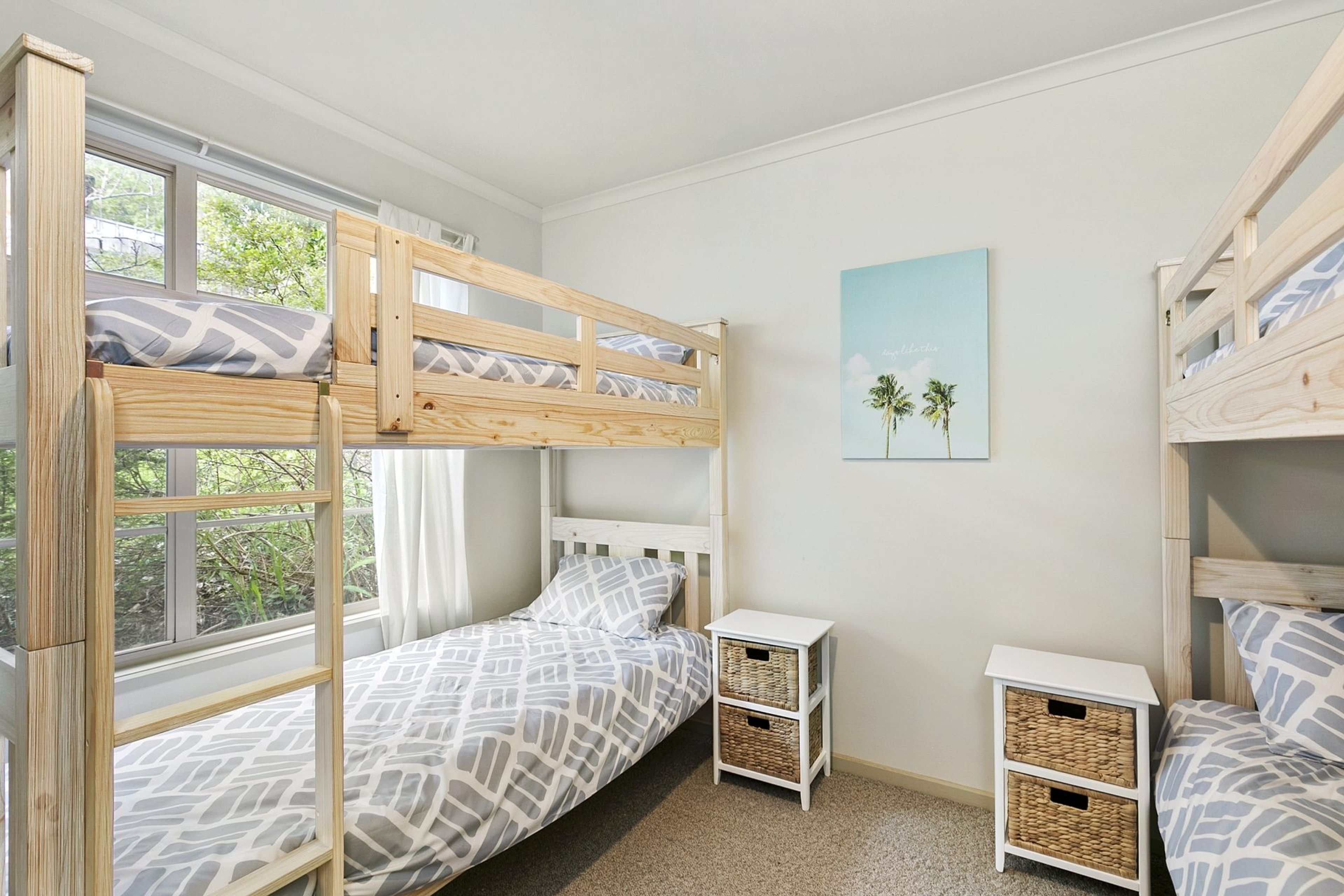 A child's bedroom with bunk beds, a small bedside table, a wall-mounted shelf, and a window with curtains.