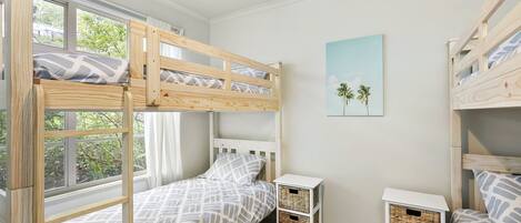 A child's bedroom with bunk beds, a small bedside table, a wall-mounted shelf, and a window with curtains.