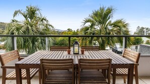 Mesa de madeira com seis cadeiras numa varanda com vista para palmeiras e céu claro.