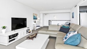 Uma sala de estar moderna com uma TV de tela plana, um rack branco, um sofá bege com almofadas azuis e uma mesa de centro branca.