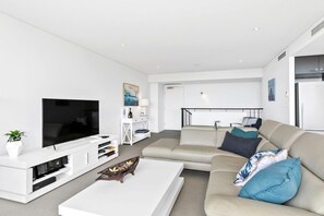 Ein modernes Wohnzimmer mit einem Flachbildfernseher, einer weißen Unterhaltungsanlage, einem beige gefärbten Sofa mit blauen Kissen und einem weißen Couchtisch.