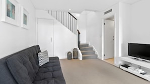 Uma sala de estar moderna com um sofá cinzento, uma TV de tela plana sobre um suporte branco, uma escada com degraus cinzas e uma porta branca.