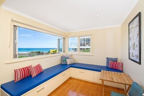 A room with a large window overlooking the sea, a built-in bench with cushions, a small table, and a framed picture on the wall. - Callahan (Marengo)