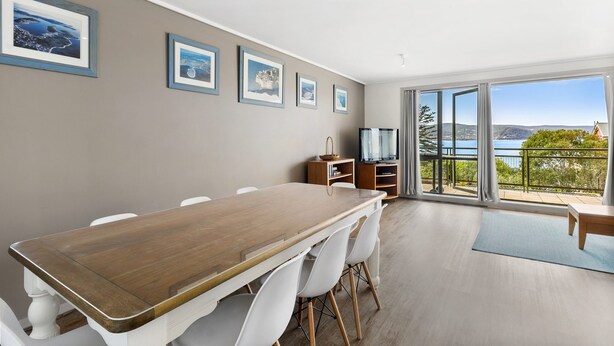 A dining area with a wooden table and white chairs, a television, and a view of the outdoors through large windows. - Apartment 29 Pacific Apartments (Lorne)