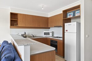 A modern kitchen with wooden cabinets, a white countertop, a microwave, a stove, and a refrigerator. - Apartment 29 Pacific Apartments (Lorne)