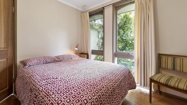 Quarto com cama, cadeira, janela com cortinas e vista para o verde lá fora.