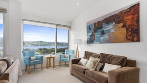 A modern living room with a large window, a brown sofa, two blue armchairs, a small table, and a painting of coastal scenery.