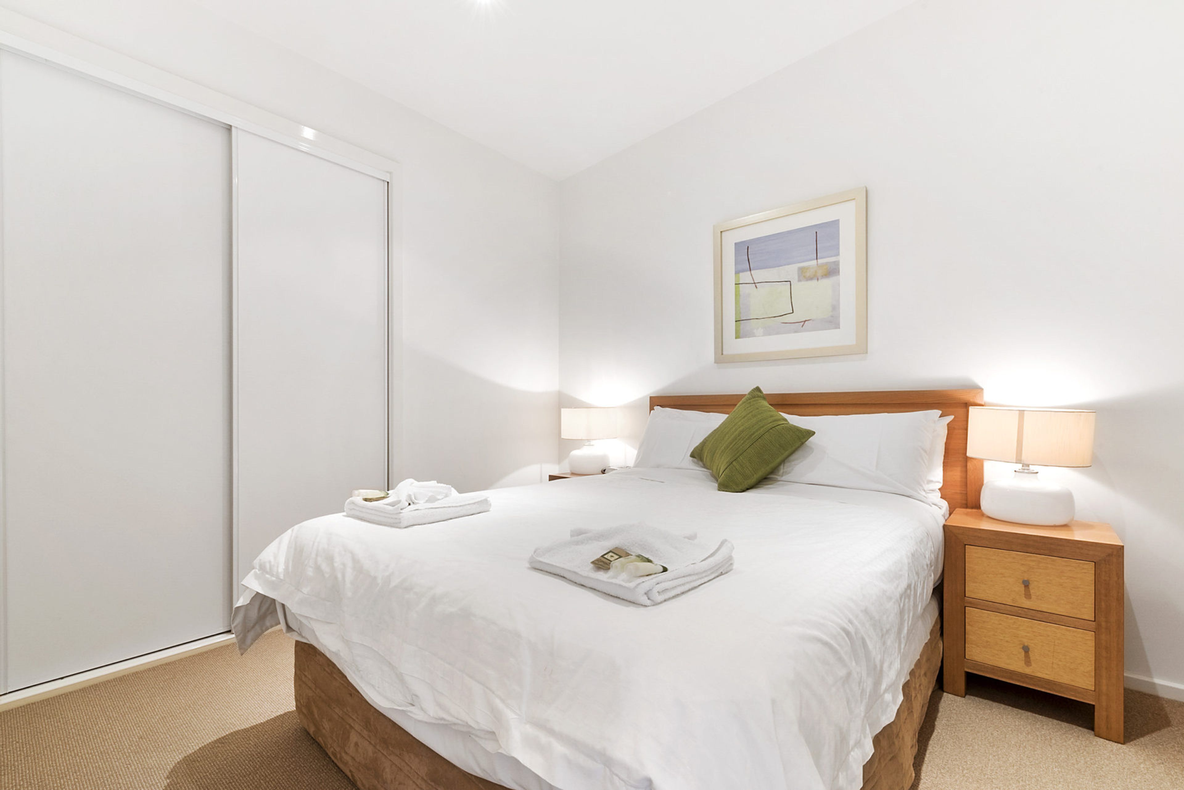 A hotel room with a bed, two bedside tables, a nightstand, a green pillow, a framed picture on the wall, and a sliding door.